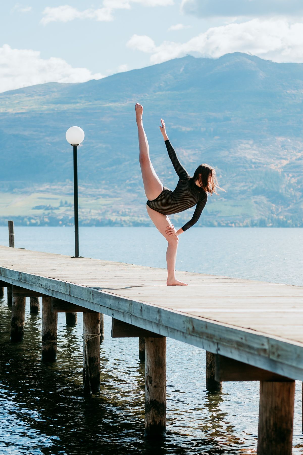 Dancer performs an elegant split pose on a wooden pier overlooking a serene mountain lake at sunset.