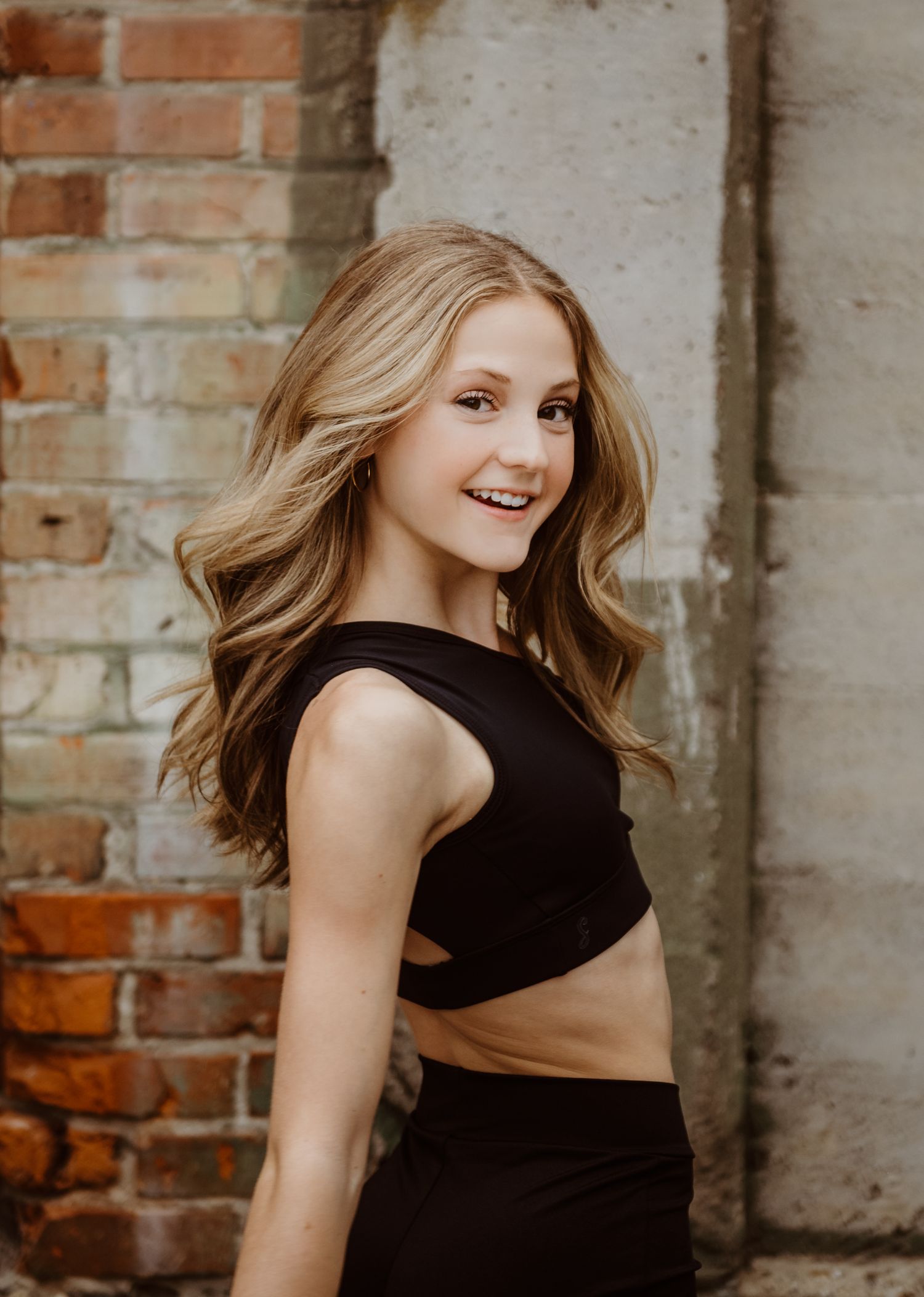 A person wearing a black crop top poses against a rustic brick wall with flowing caramel-colored wavy hair.