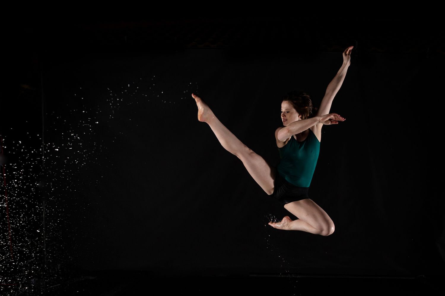 Dancer in teal leotard performs graceful leap against black background with water splashing effect.