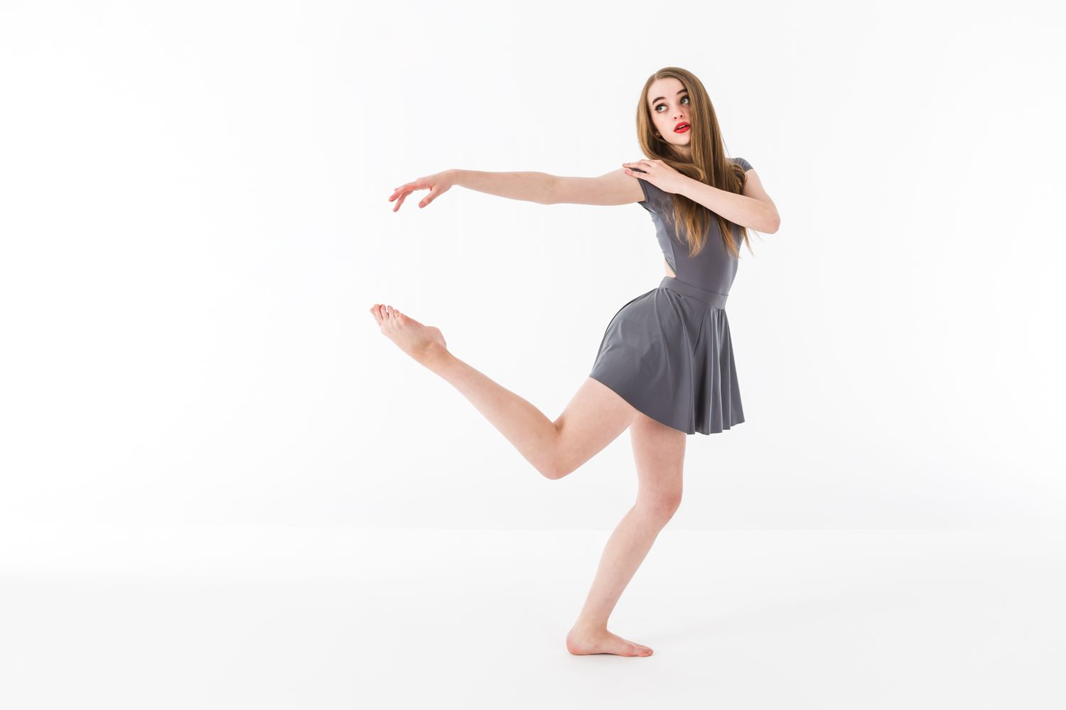 Dancer in grey dress performs elegant pose with extended leg and graceful arm movement.