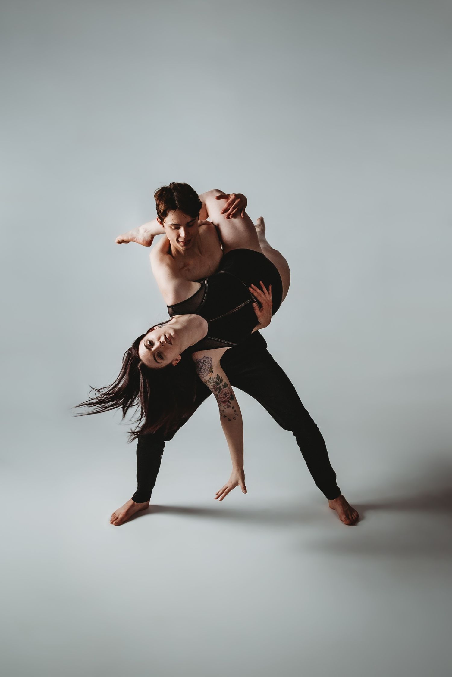 Two dancers perform intimate contemporary dance lift in minimalist studio setting.