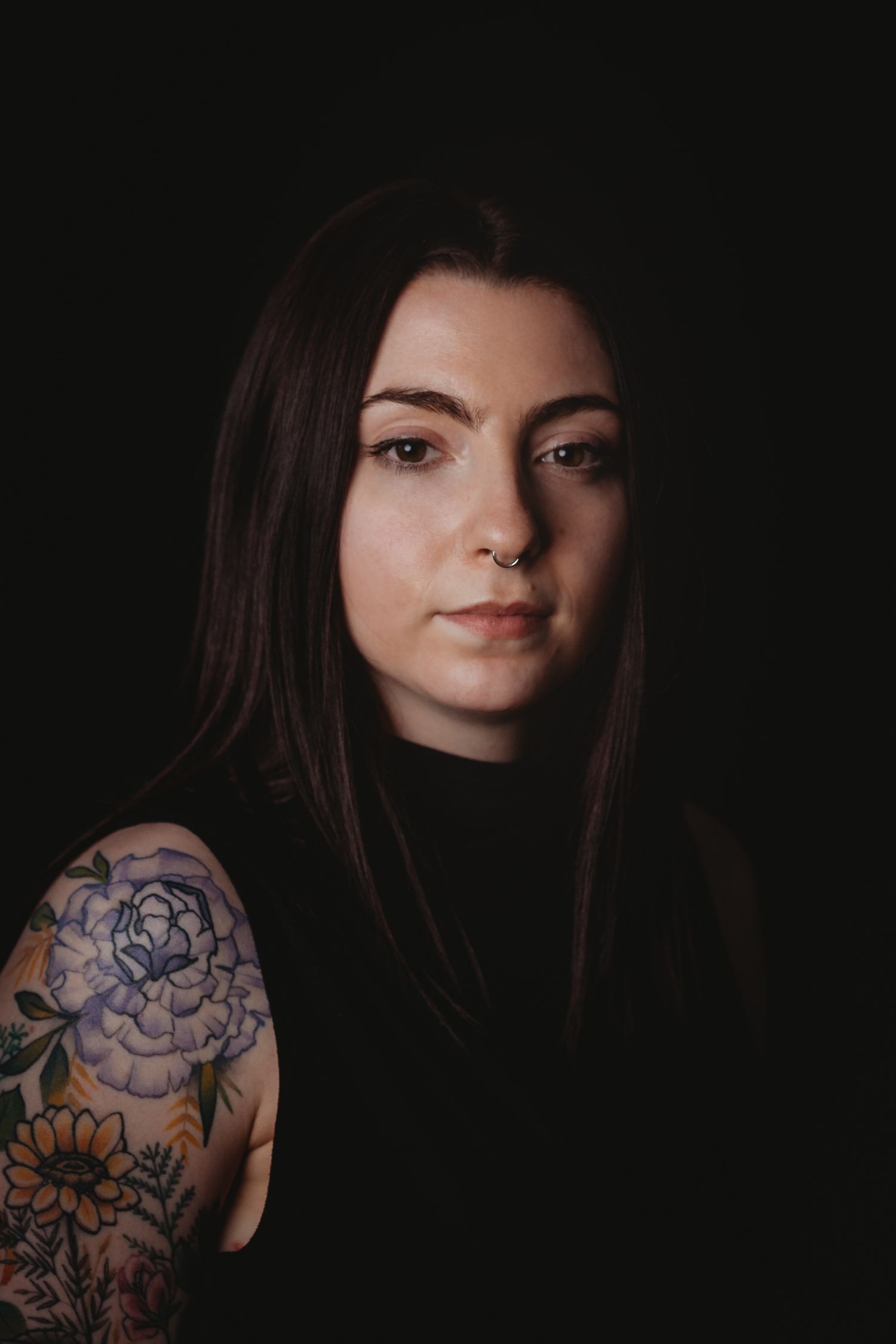 Portrait of someone with dark hair wearing black and featuring a floral arm tattoo against a dark backdrop.