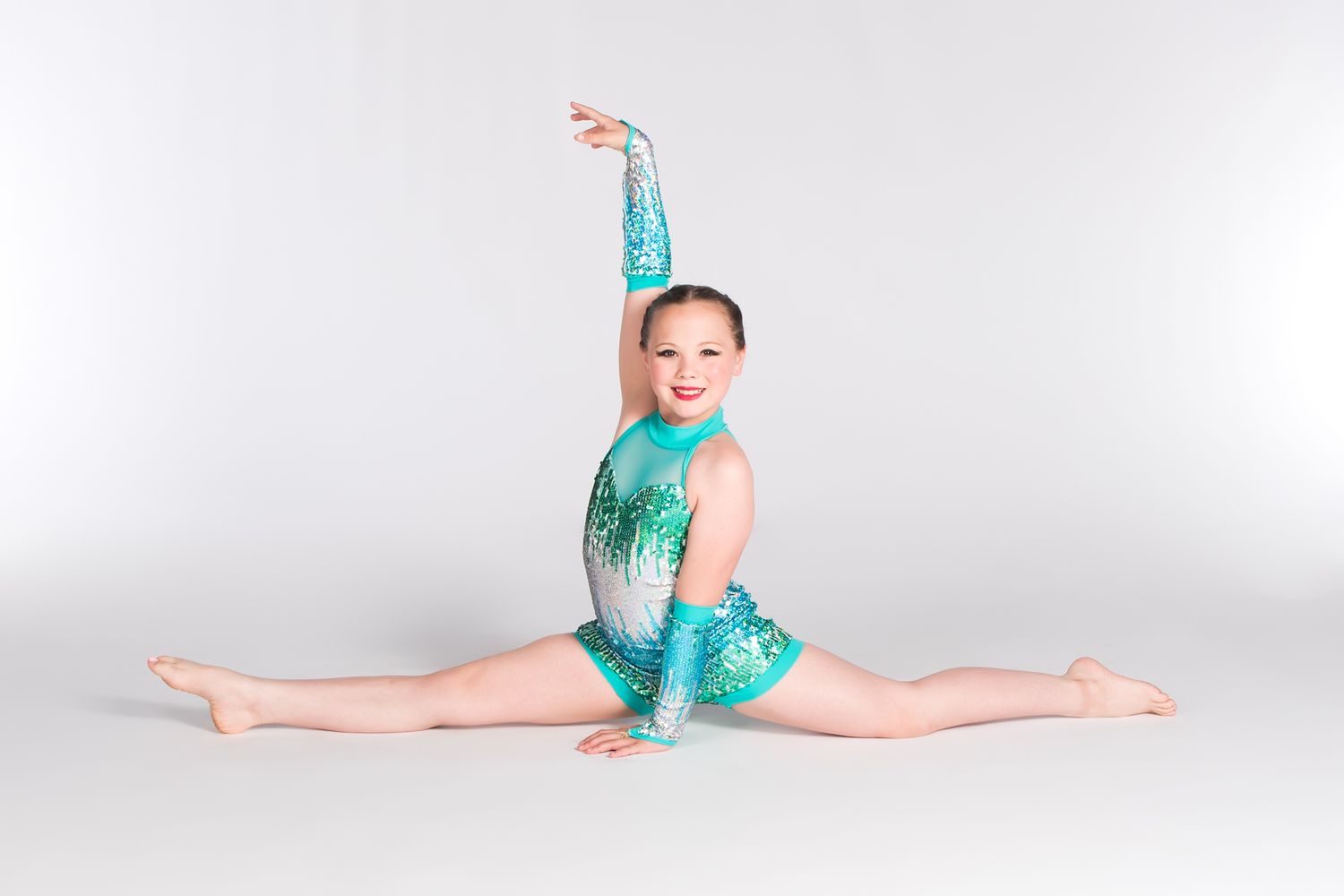 Gymnast in sparkly turquoise leotard performs splits pose with raised arm on white background.