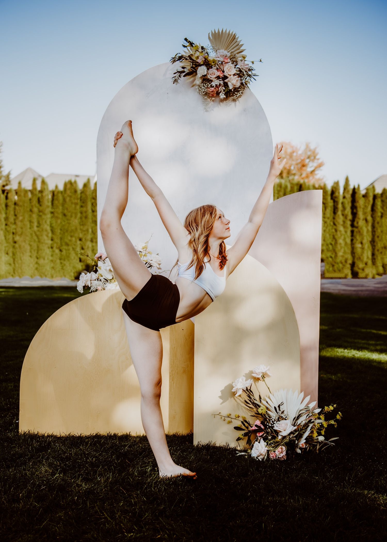Elegant dancer in black leotard strikes graceful pose against curved white and cream backdrop with floral arrangements outdoors.
