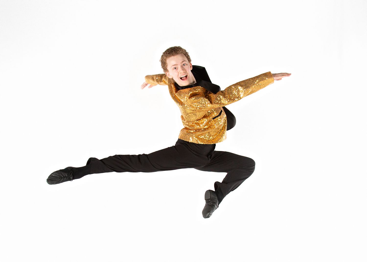 A dancer in a gold top and black pants performs an athletic leap against a white background.