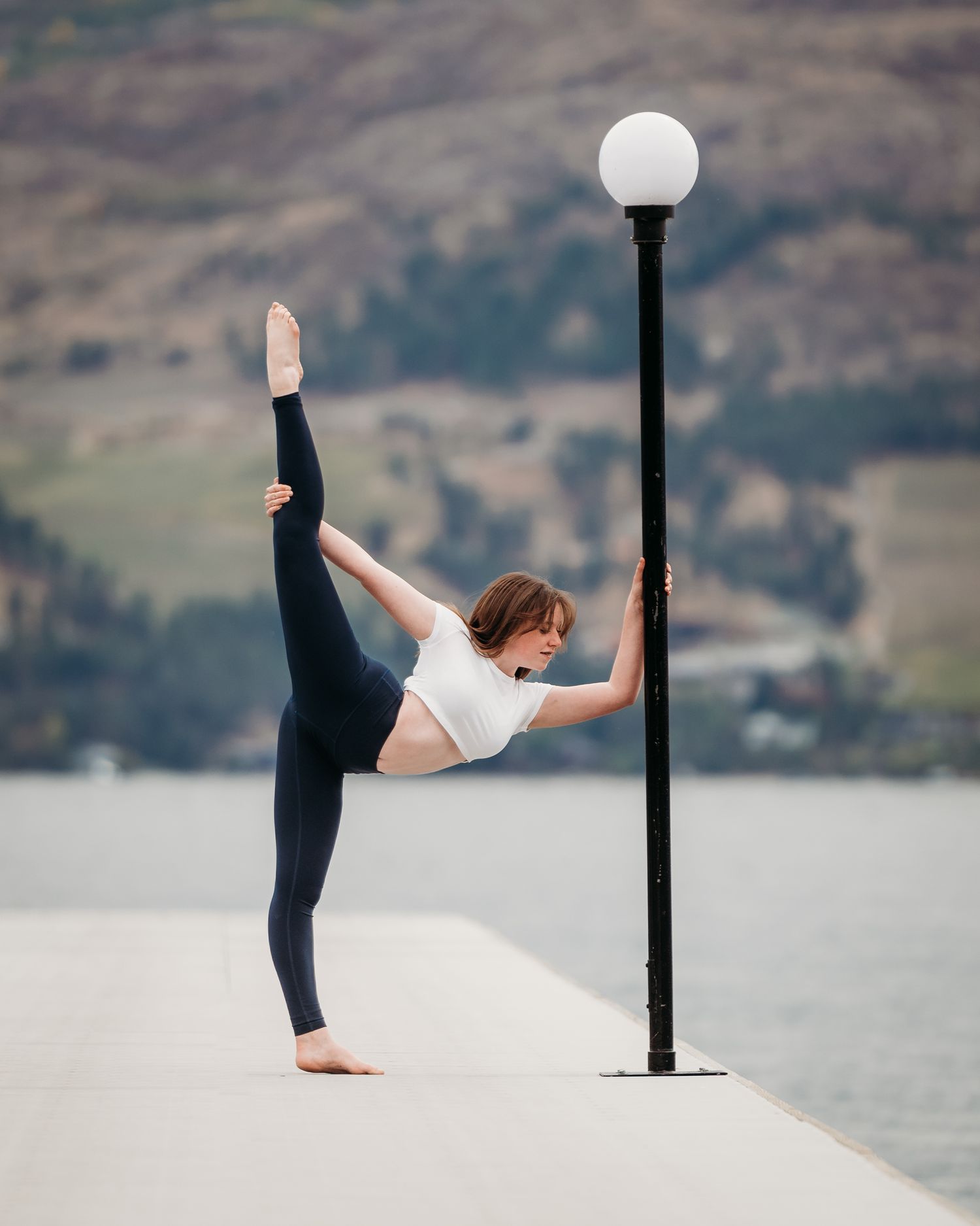 A dancer performs an elegant split pose against a lakeside lamp post, showcasing flexibility in athletic wear.