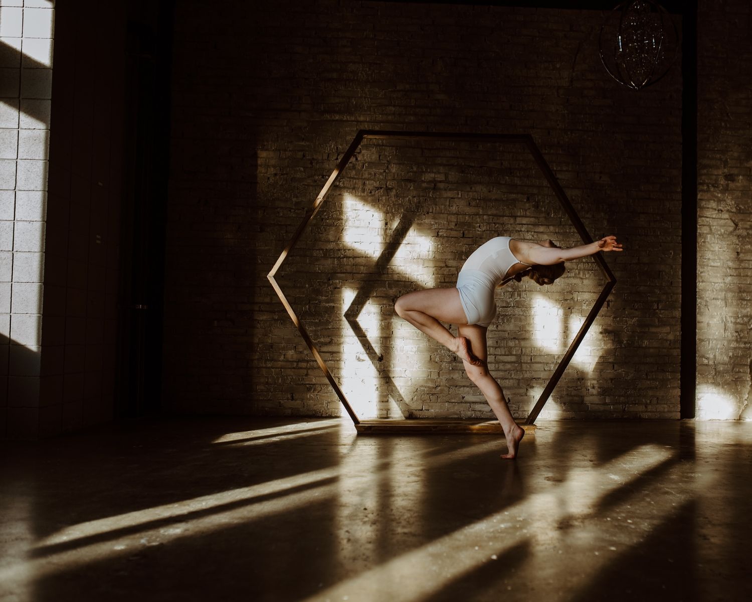 Graceful dancer poses in dramatic lighting against a geometric hexagonal light fixture casting shadows on dark walls.