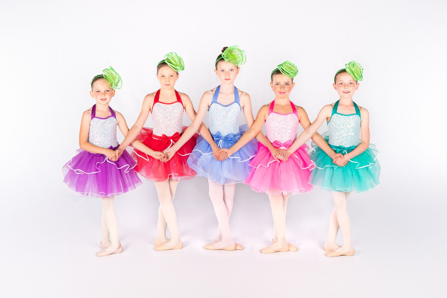 Five young ballet dancers wearing colorful tutus and matching headdresses pose together in ballet positions against white background.