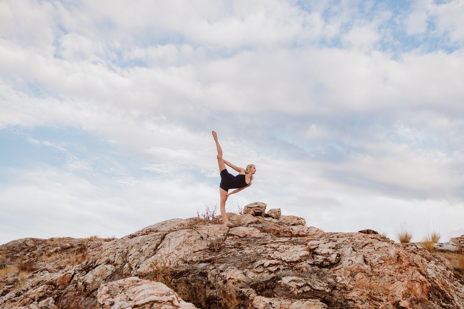 A person performs an elegant dance pose atop a rocky hill against a cloudy sky backdrop.
