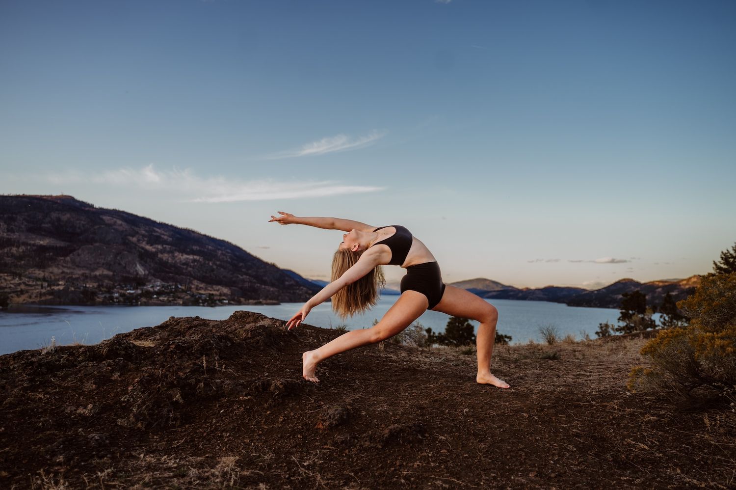 Dancer performs graceful leap pose against scenic mountain lake backdrop on rocky terrain at sunset.