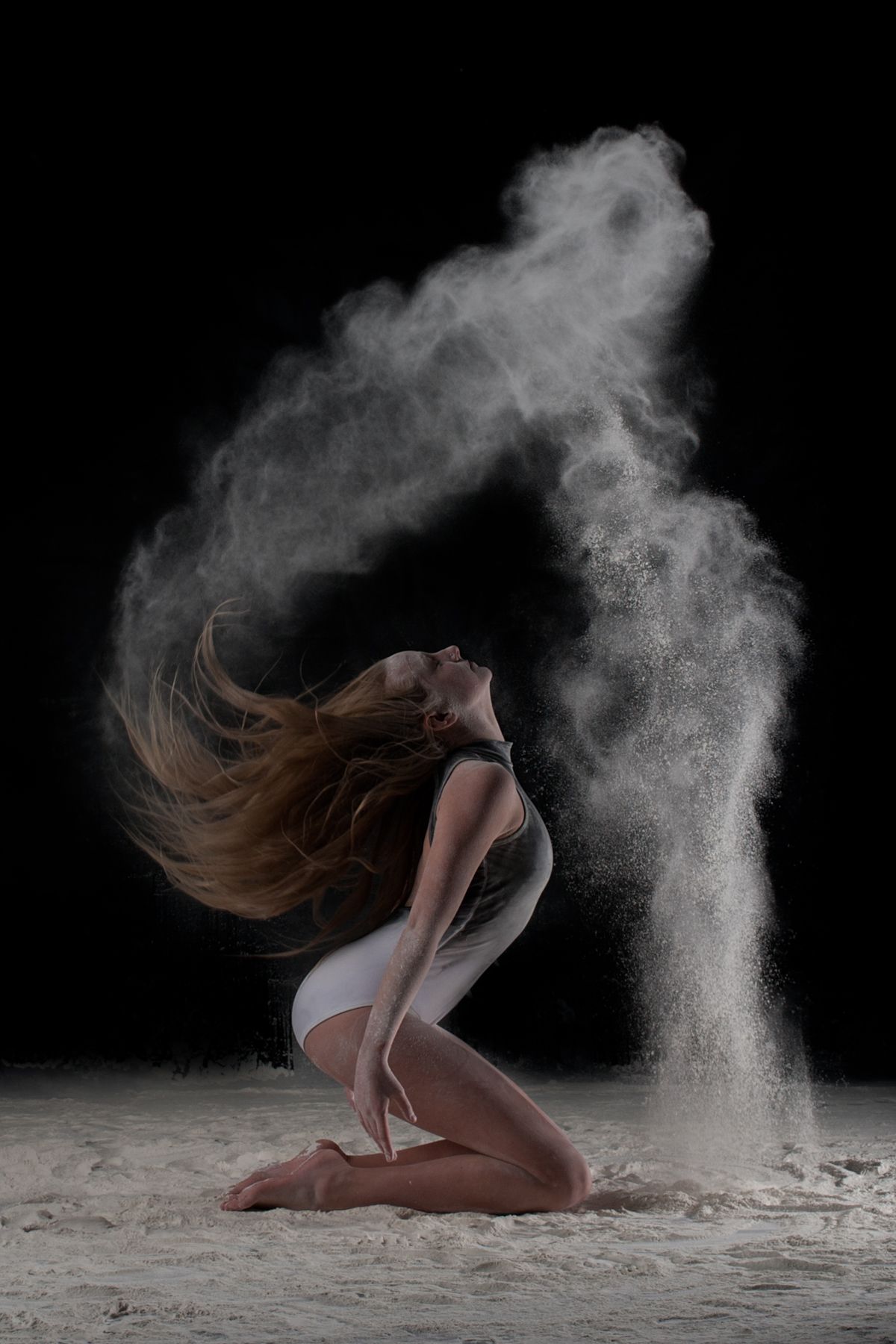 Dancer kneeling in flour dust creating dynamic motion effect with hair tossed back on dark background.