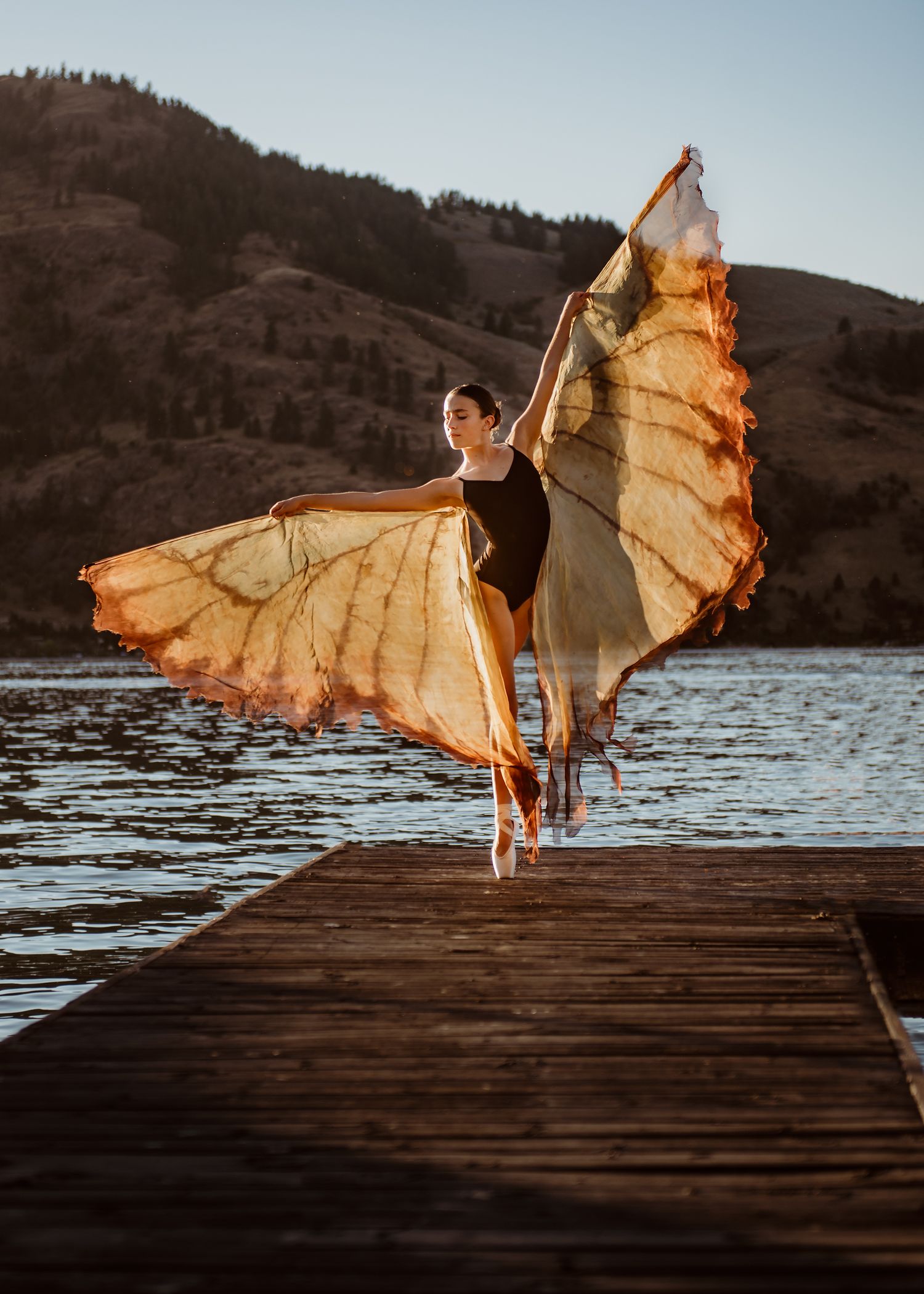 Ballet dancer poses gracefully on lakeside dock at sunset with flowing golden fabric wings creating ethereal effect.