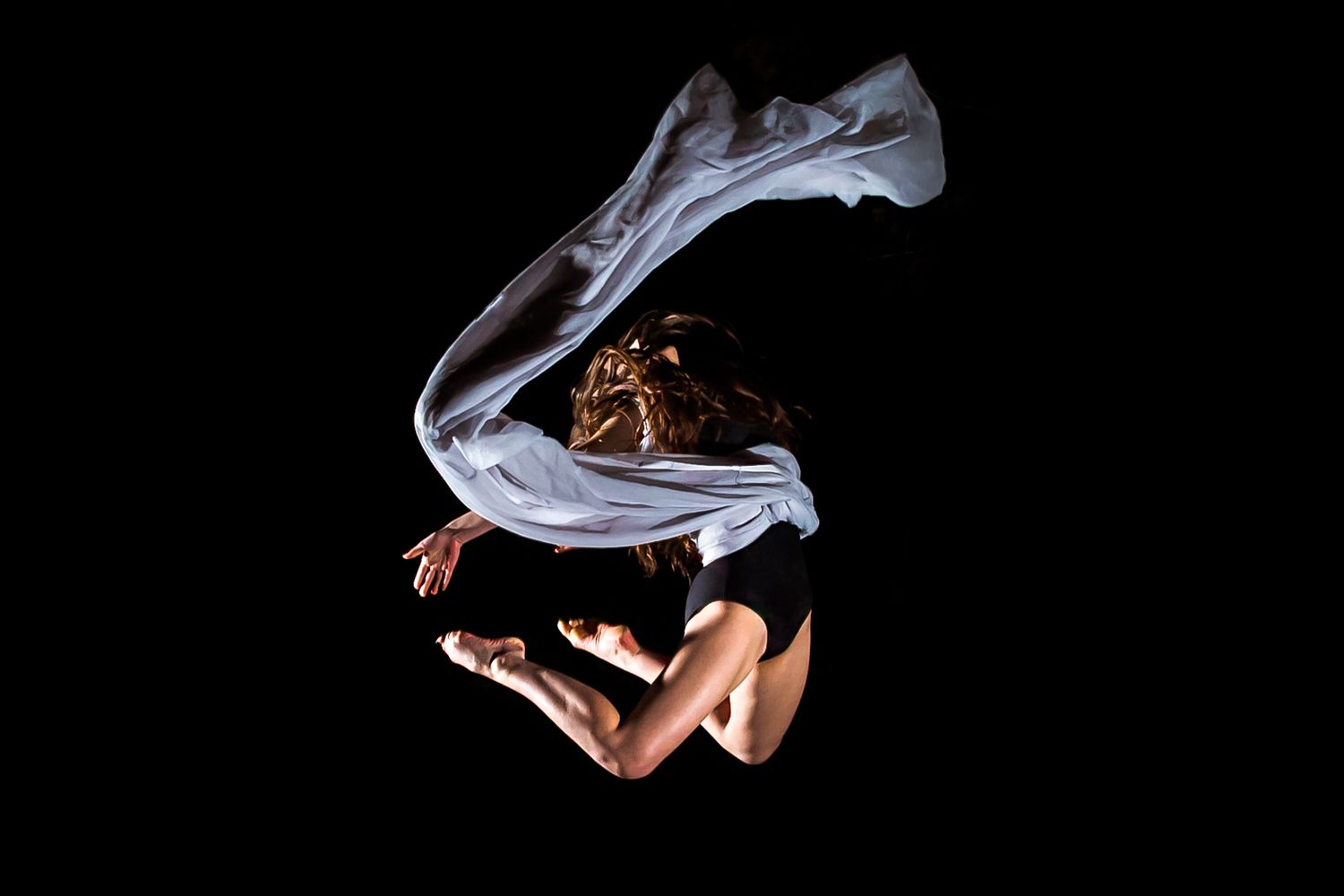 Athletic dancer in flowing white fabric performs a dynamic aerial pose against black background.