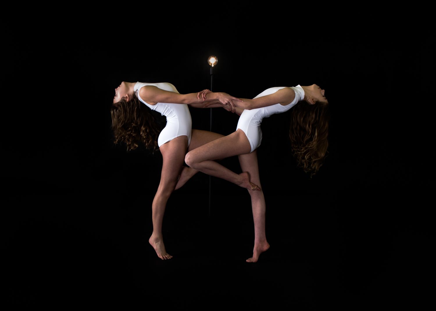 Two gymnasts perform a synchronized aerial pose against a black background, creating a mirror-like artistic formation.