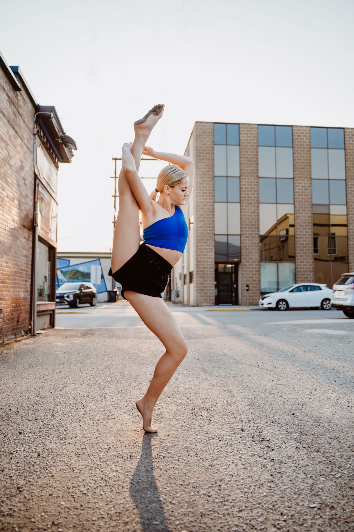 A dancer in blue and black athletic wear performs a graceful high kick on a street in front of brick buildings.