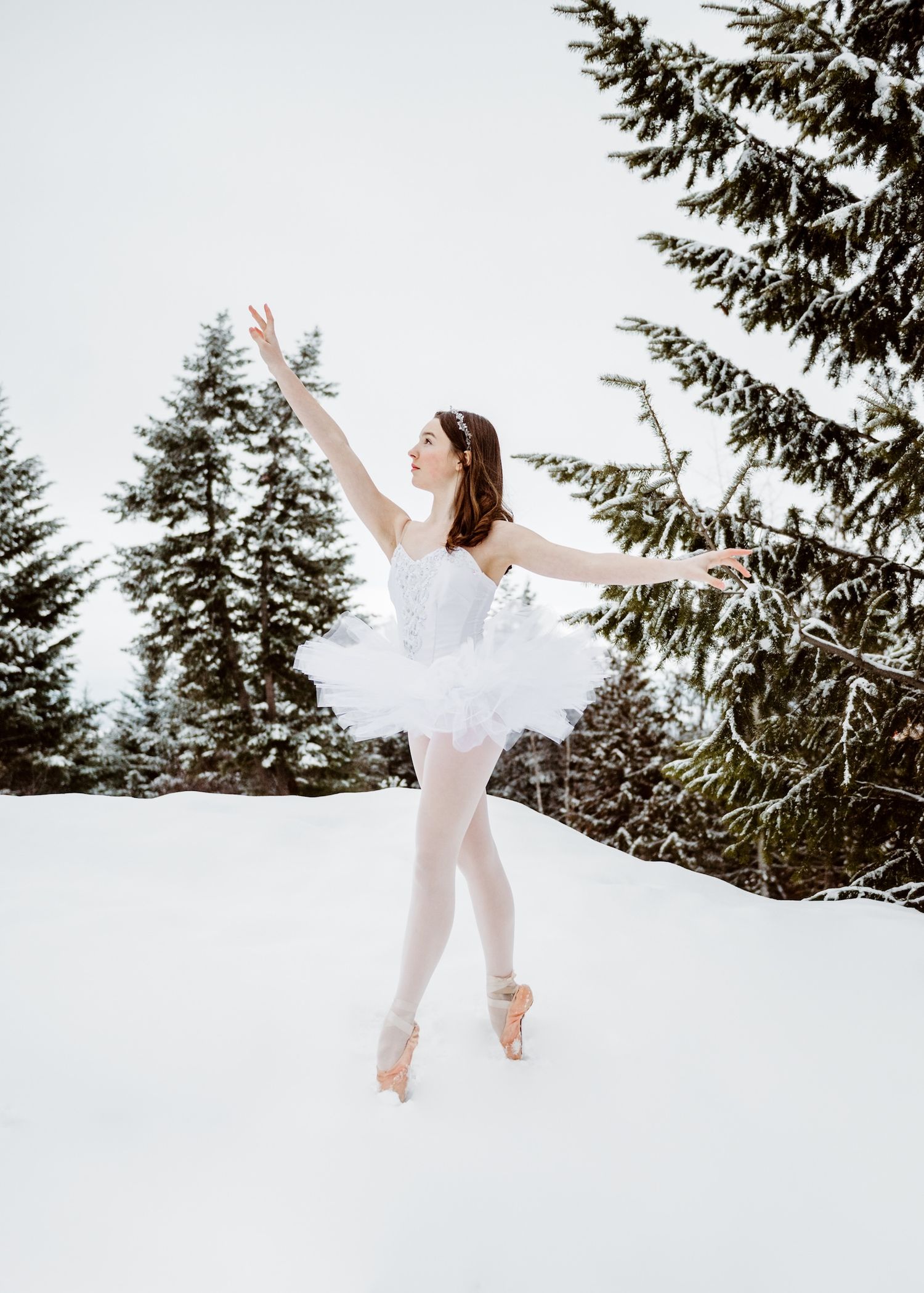 A ballet dancer in white performs an elegant pose on snowy ground surrounded by evergreen trees in winter.