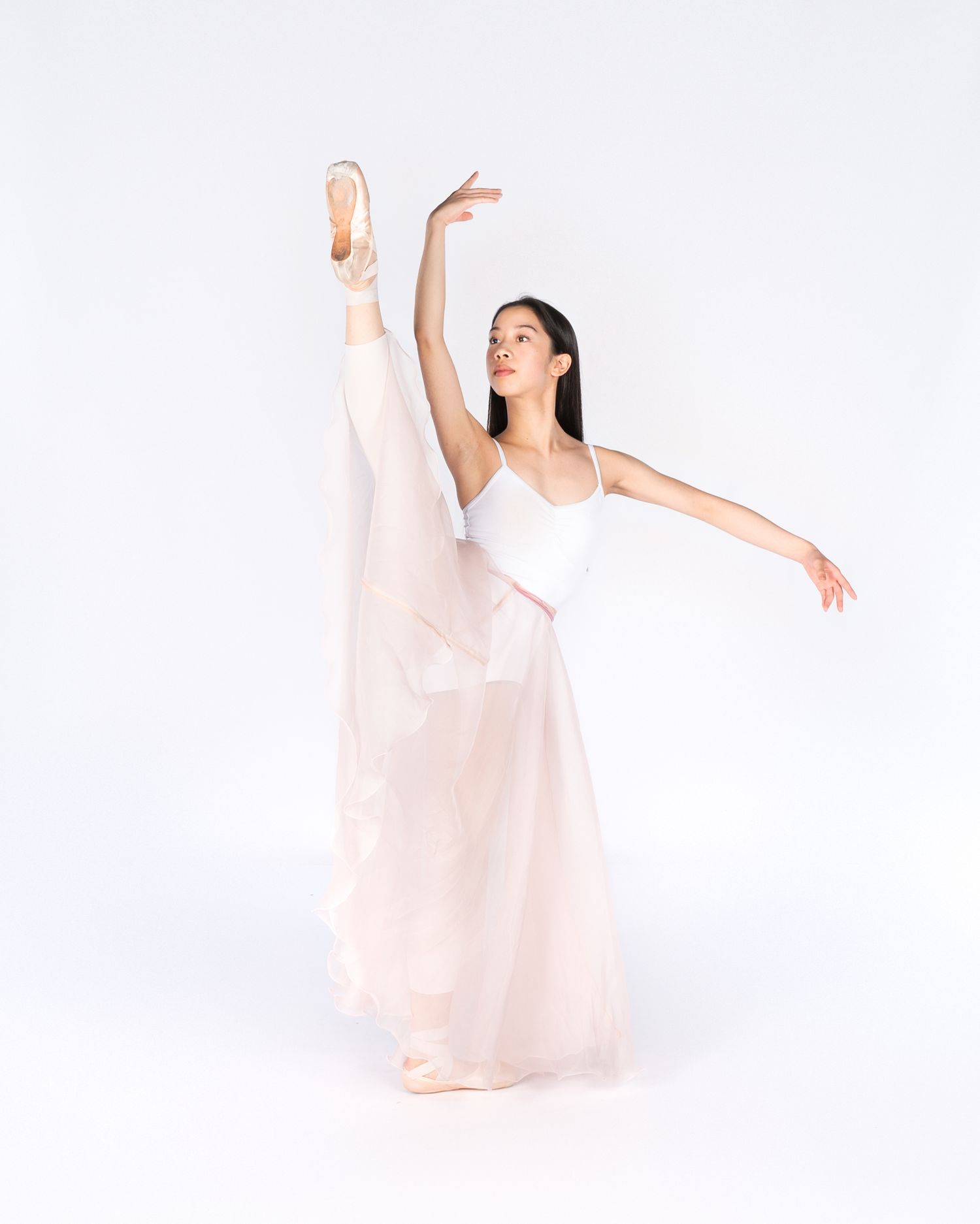 Ballet dancer in flowing white dress performs elegant arabesque pose against white backdrop.