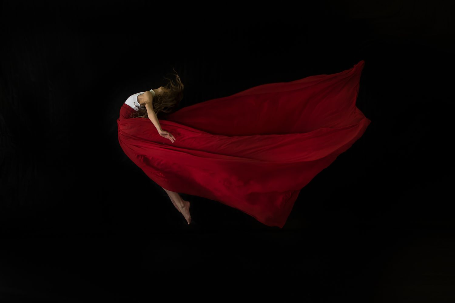 Figure in flowing red fabric floats gracefully against a dark background in dramatic artistic photography.
