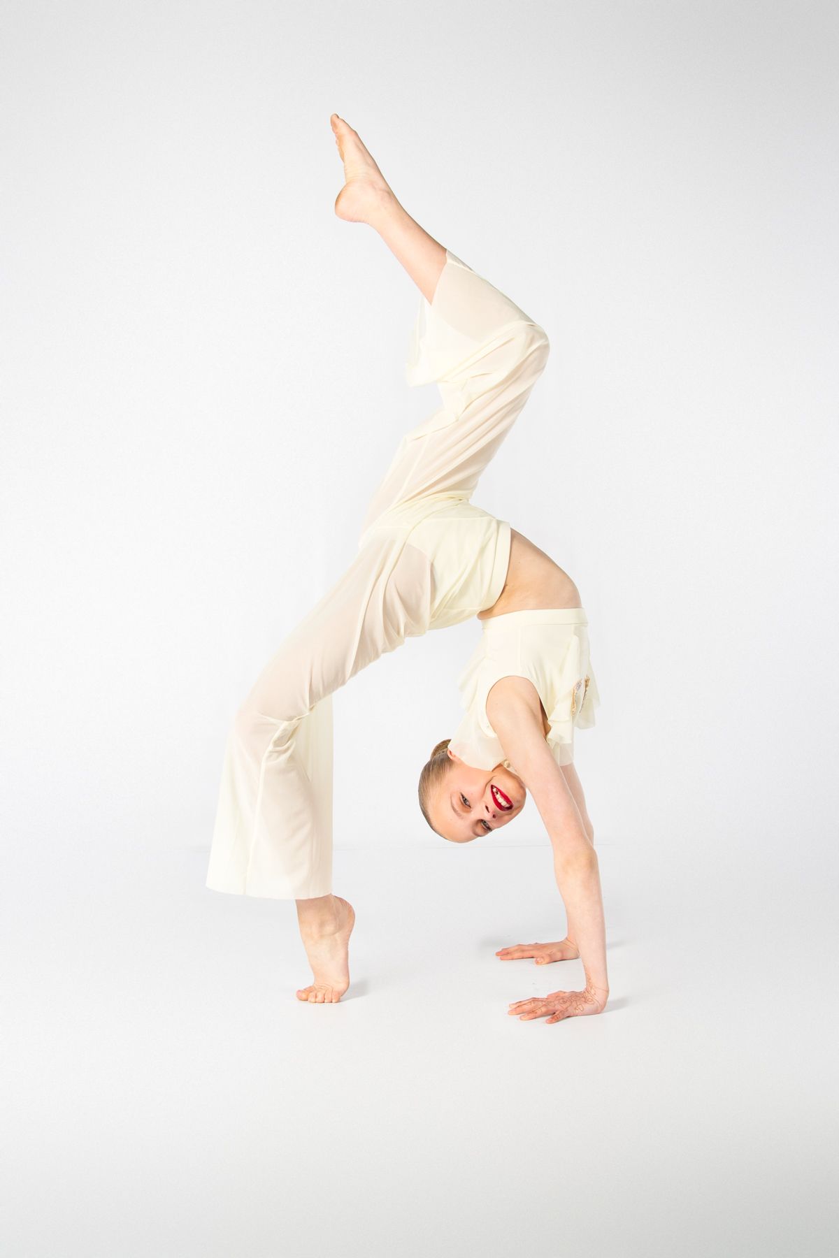 Modern dancer in flowing white attire performs a graceful backbend bridge pose against a light studio background.