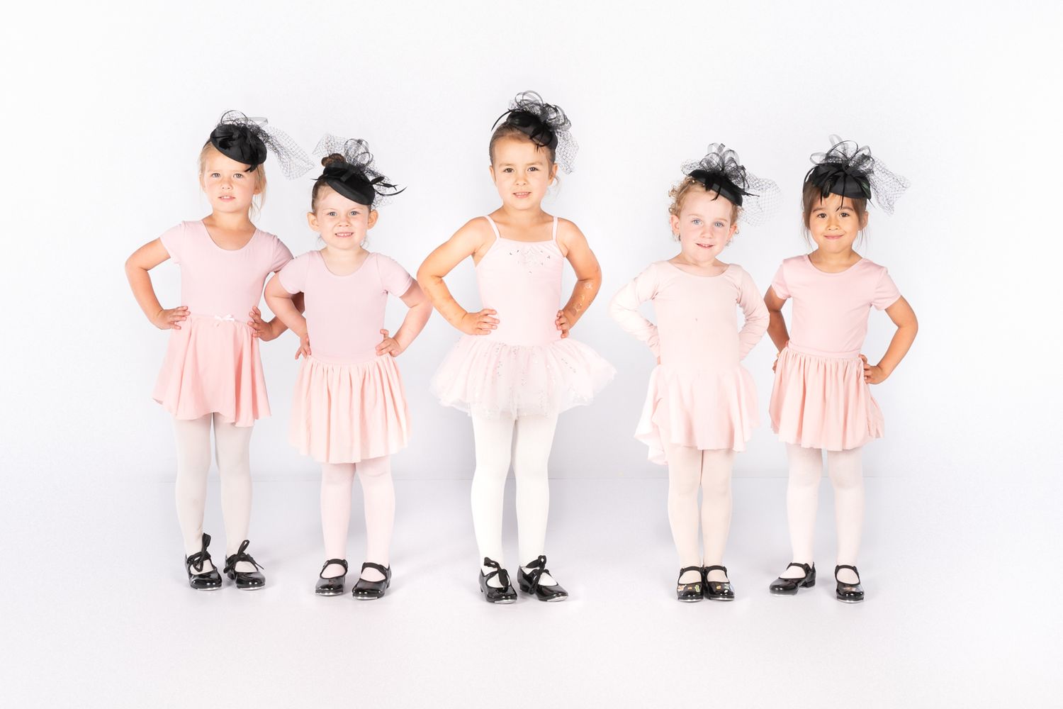 Five young dancers in pink outfits and tap shoes pose in a line against a white backdrop.