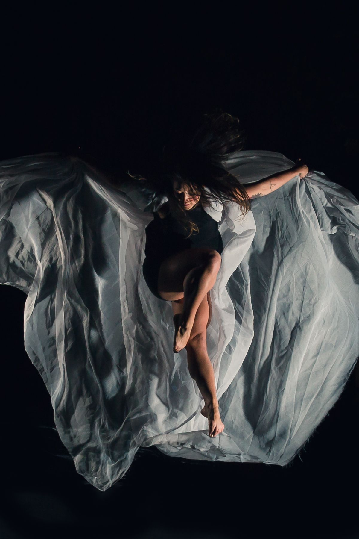 A dancer in black performs an elegant pose against billowing white fabric on a dark background.