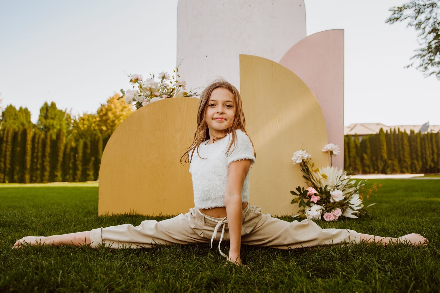 A person in white clothing does a full split pose on grass in front of a modern architectural backdrop with golden details.