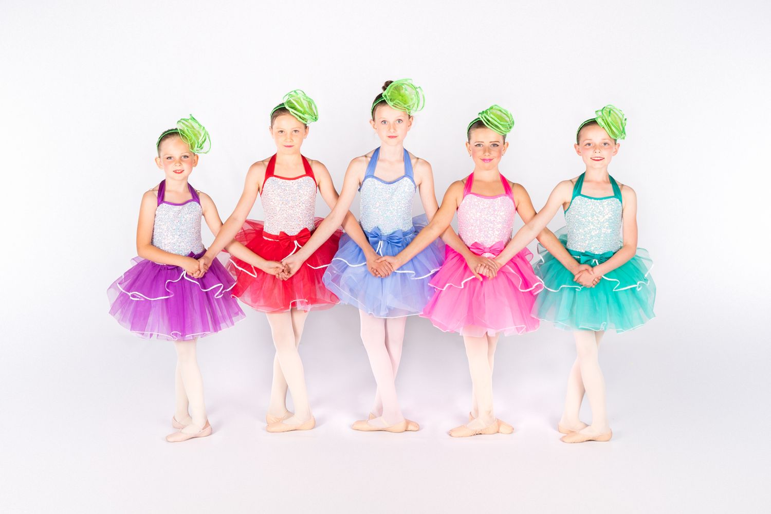 Five young ballet dancers in colorful tutus and green headbands pose in first position.