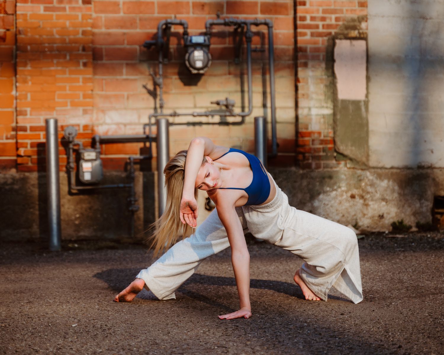 Dancer in white pants and blue top performs dramatic pose against industrial brick wall backdrop with exposed pipes.