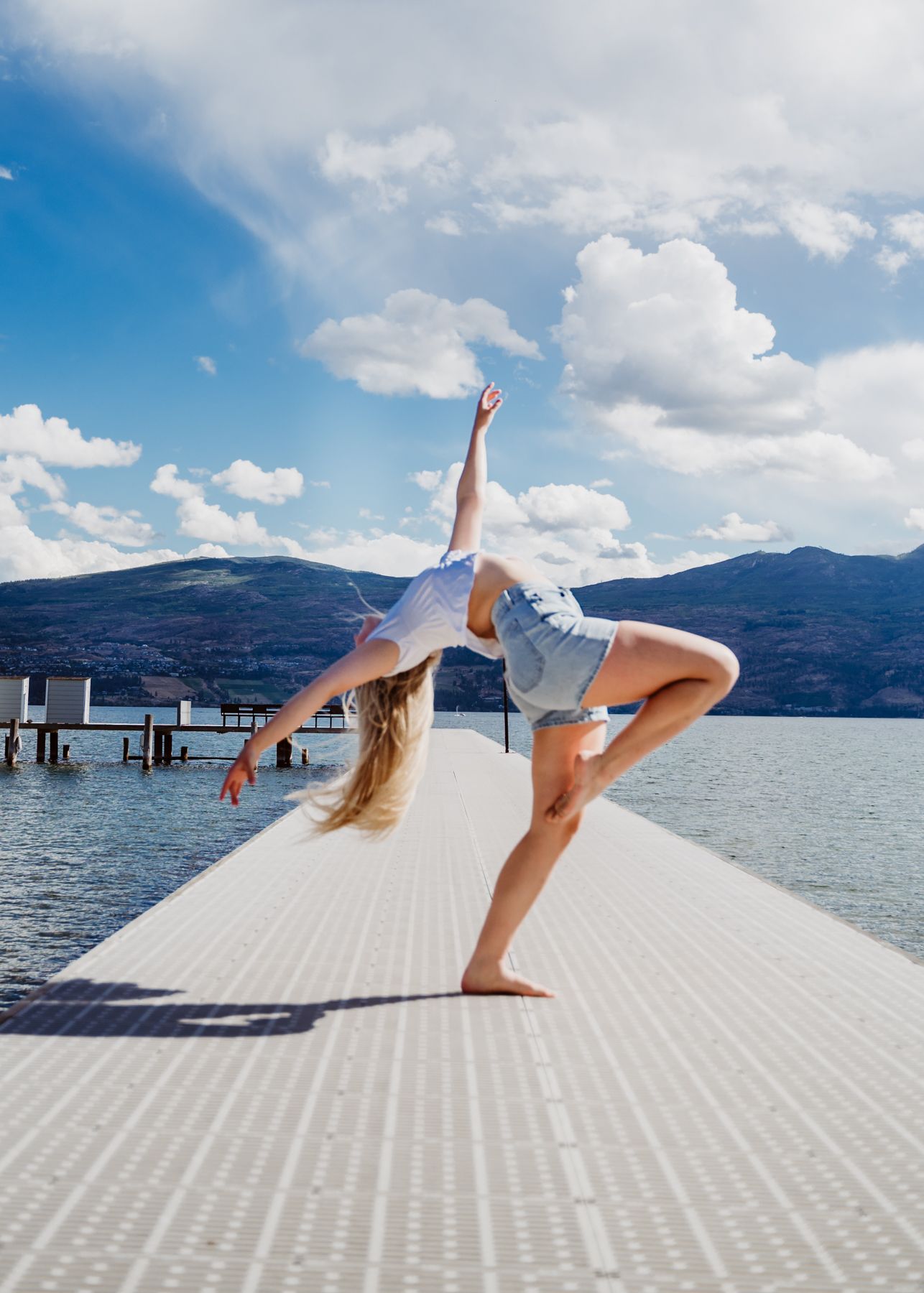 Dancer performs a backbend on a dock by the lake with mountains and fluffy clouds in the background.