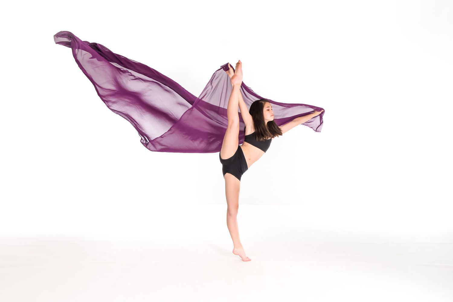 Graceful dancer in black leotard strikes elegant pose with flowing purple fabric against white background.