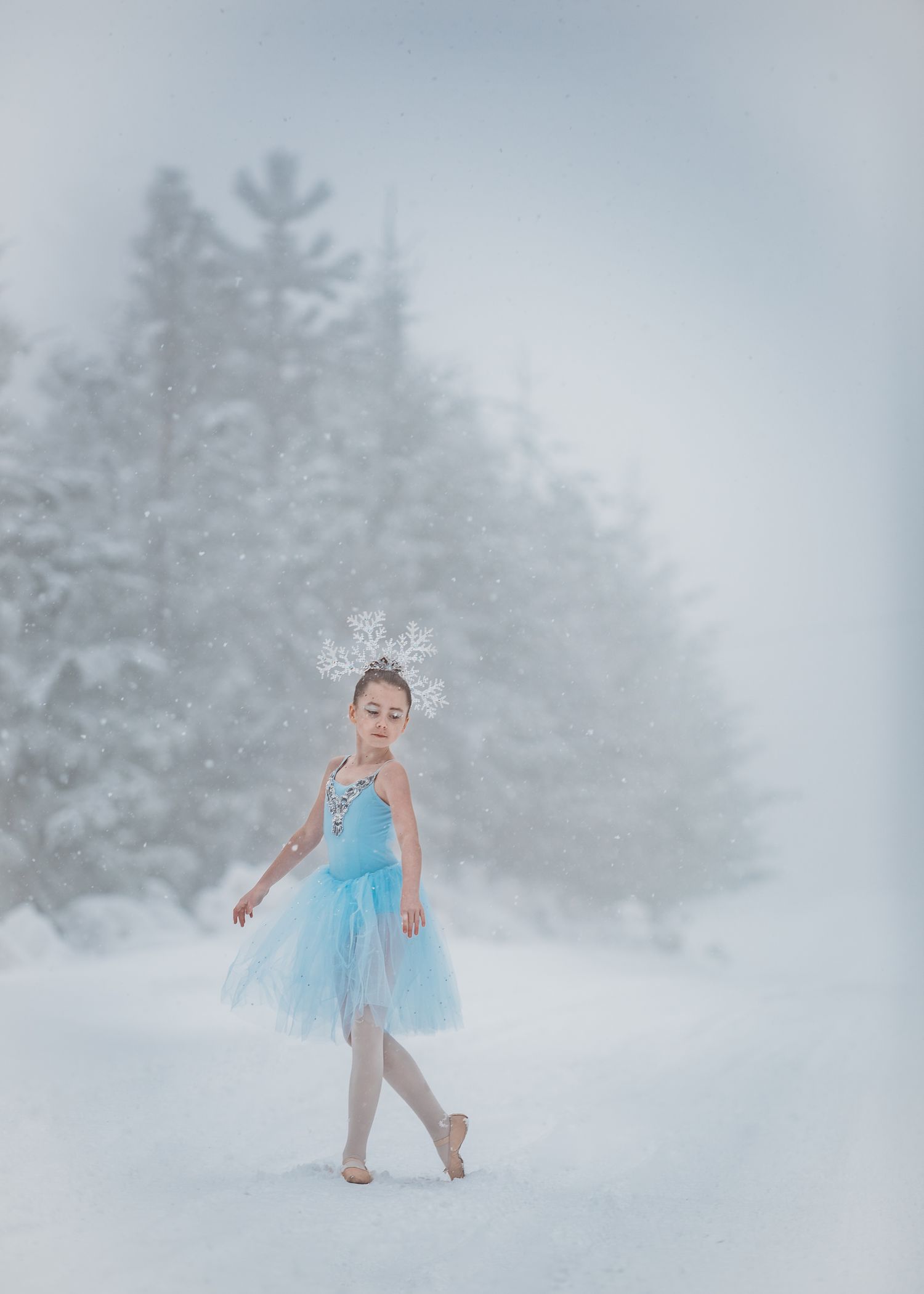 A ballet dancer in a light blue tutu dress and crown poses gracefully in a snowy winter forest setting.