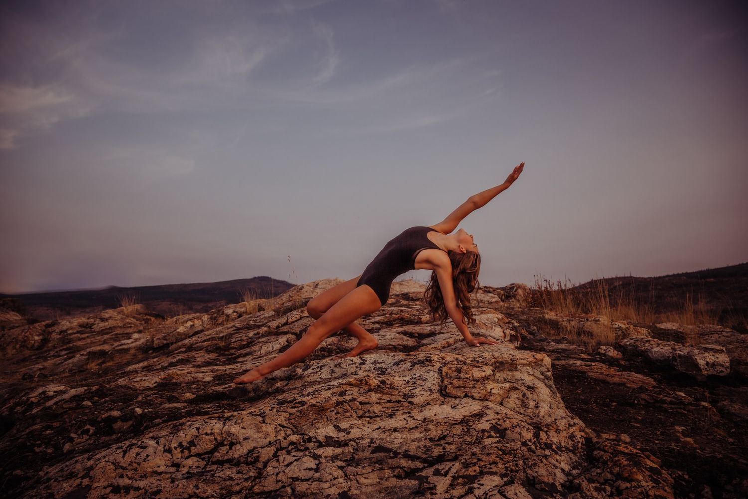 Athletic dancer stretches in artistic pose on rocky cliff during purple dusk sky.