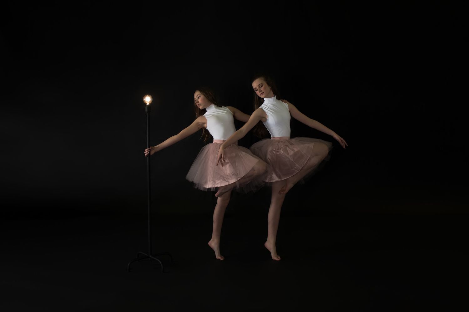 Two ballet dancers in pink tutus perform synchronized poses against a dark background while holding a glowing light.