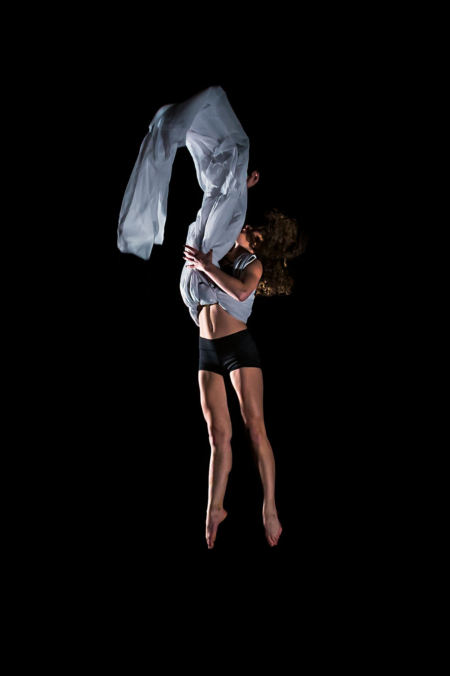 A dancer leaps gracefully against a black background wearing a flowing white top and black shorts.