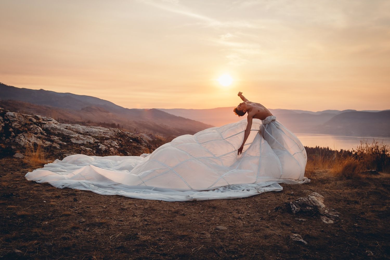 A flowing white wedding dress billows in the wind against a golden sunset over mountain peaks in a dramatic landscape.