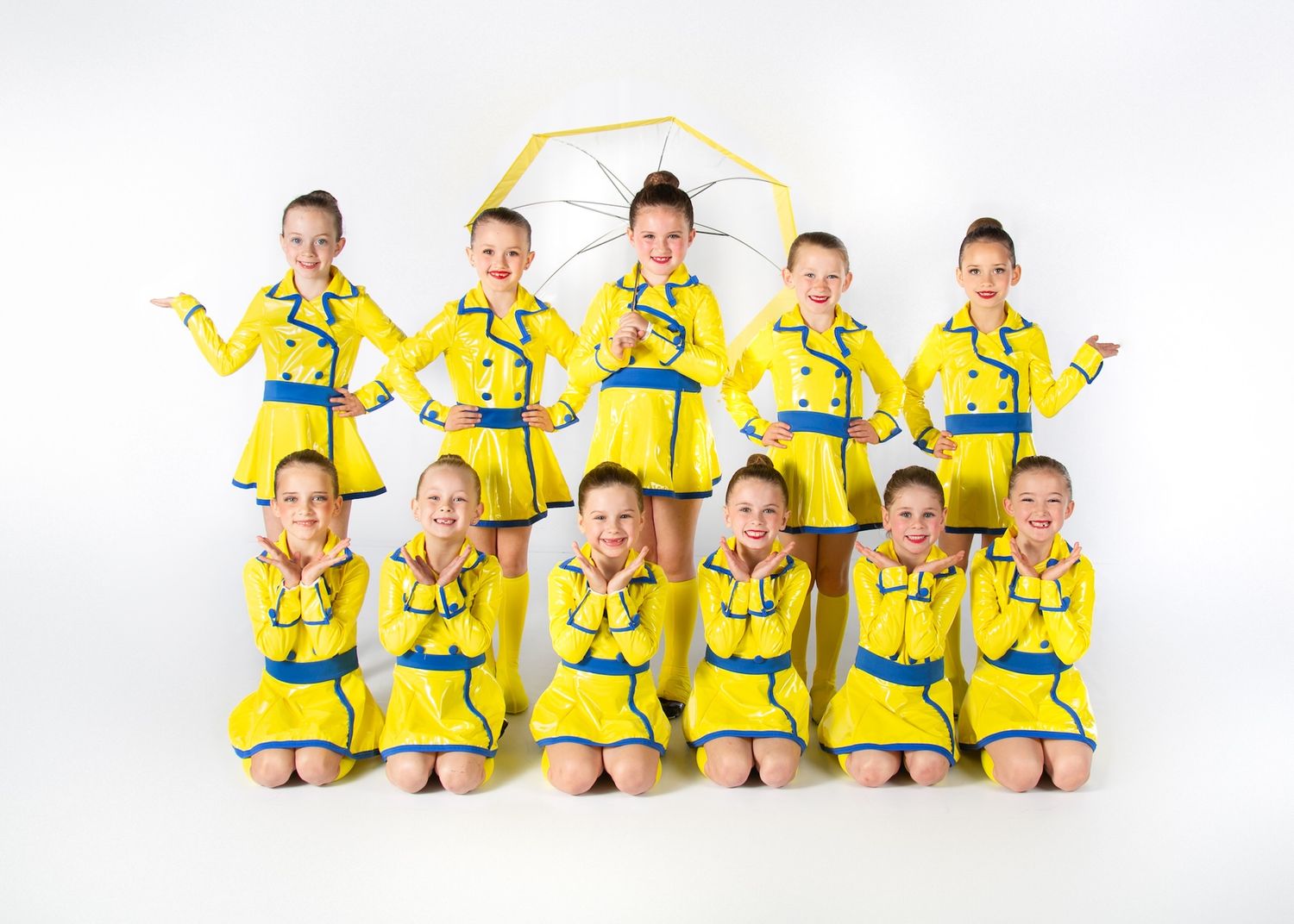 Dance group in bright yellow raincoats and blue belts posing in two rows on white background.