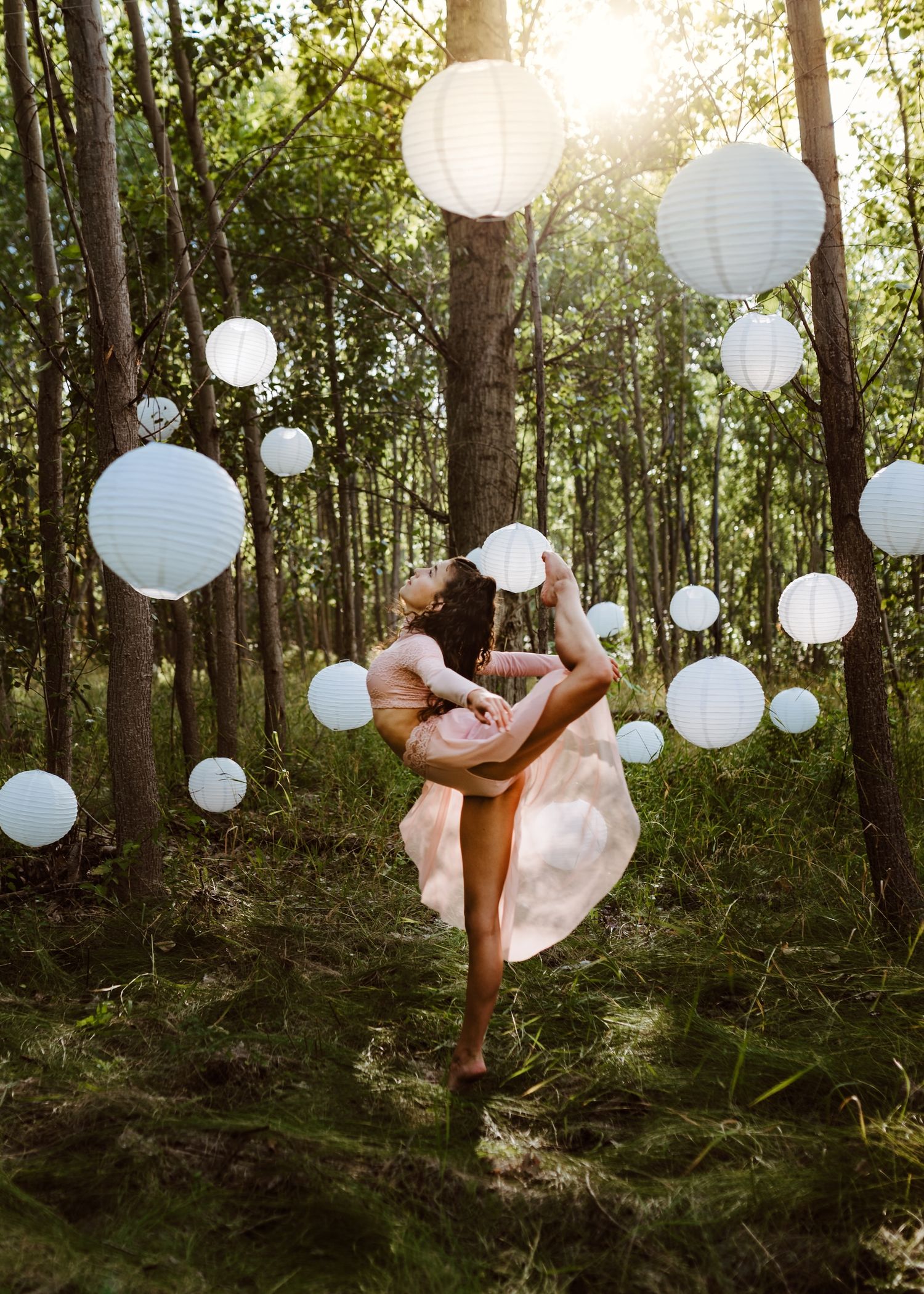 Dancer in flowing dress performs in sunlit forest with hanging white paper lanterns creating magical atmosphere.