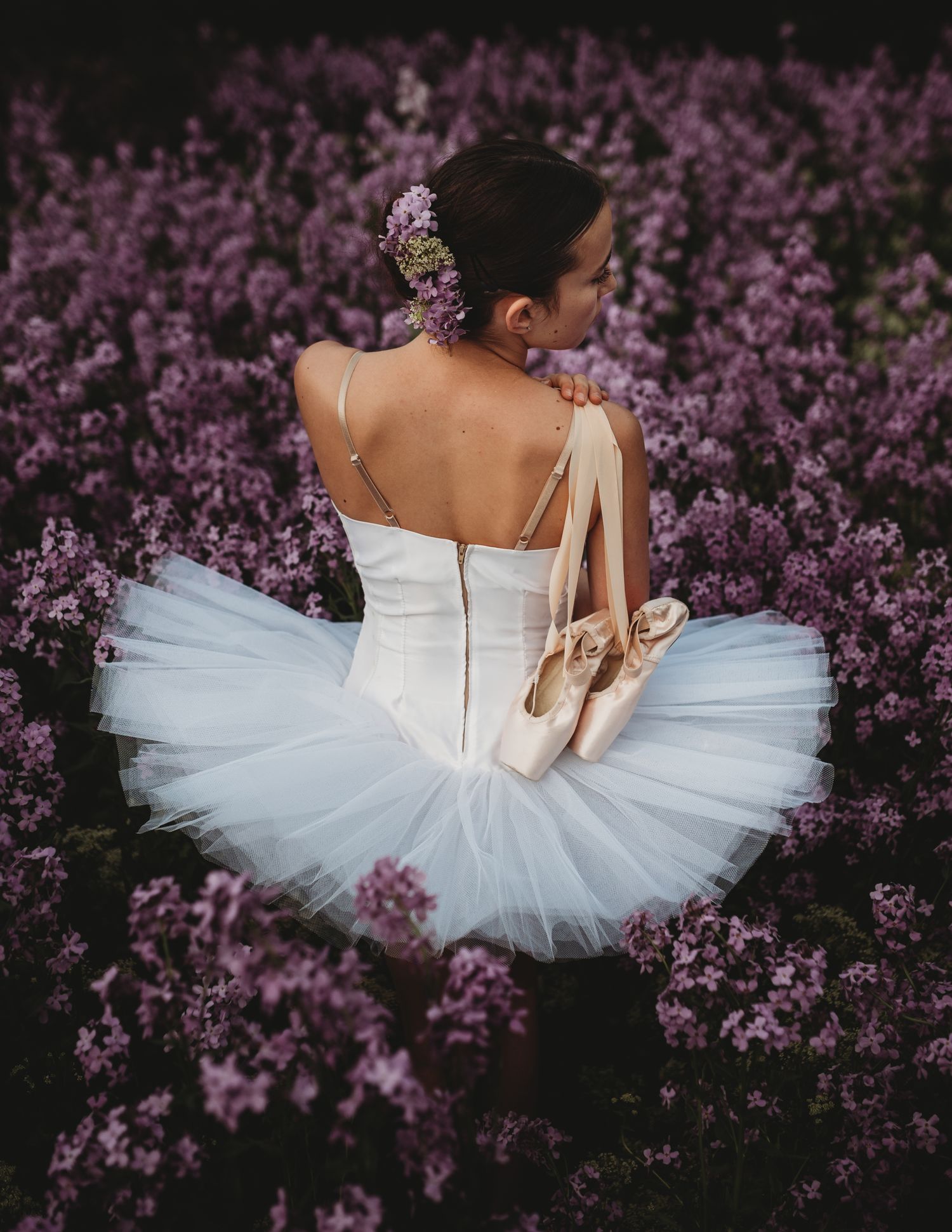 Ballet dancer in white tutu poses gracefully among blooming purple flowers in romantic outdoor setting.