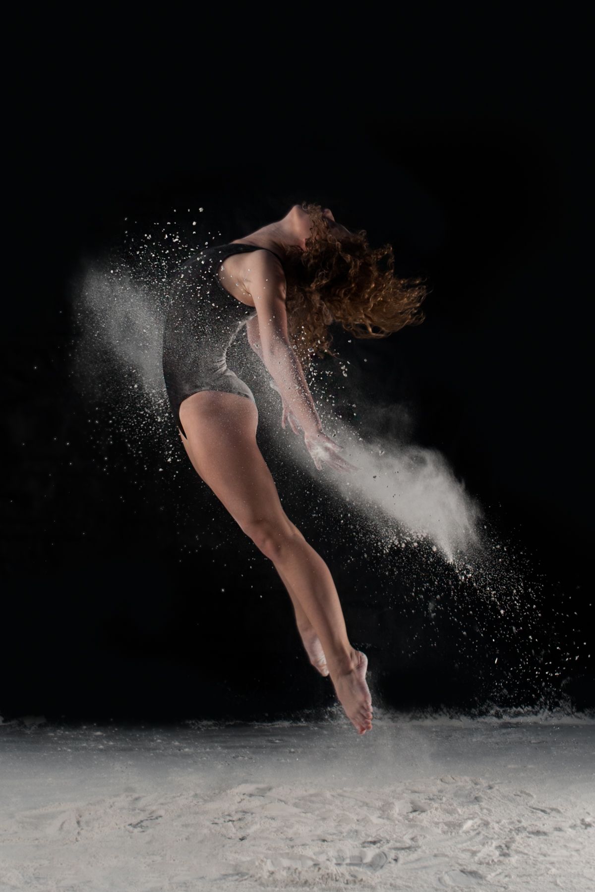 Dancer leaps gracefully through white powder creating dynamic motion effect against black background.