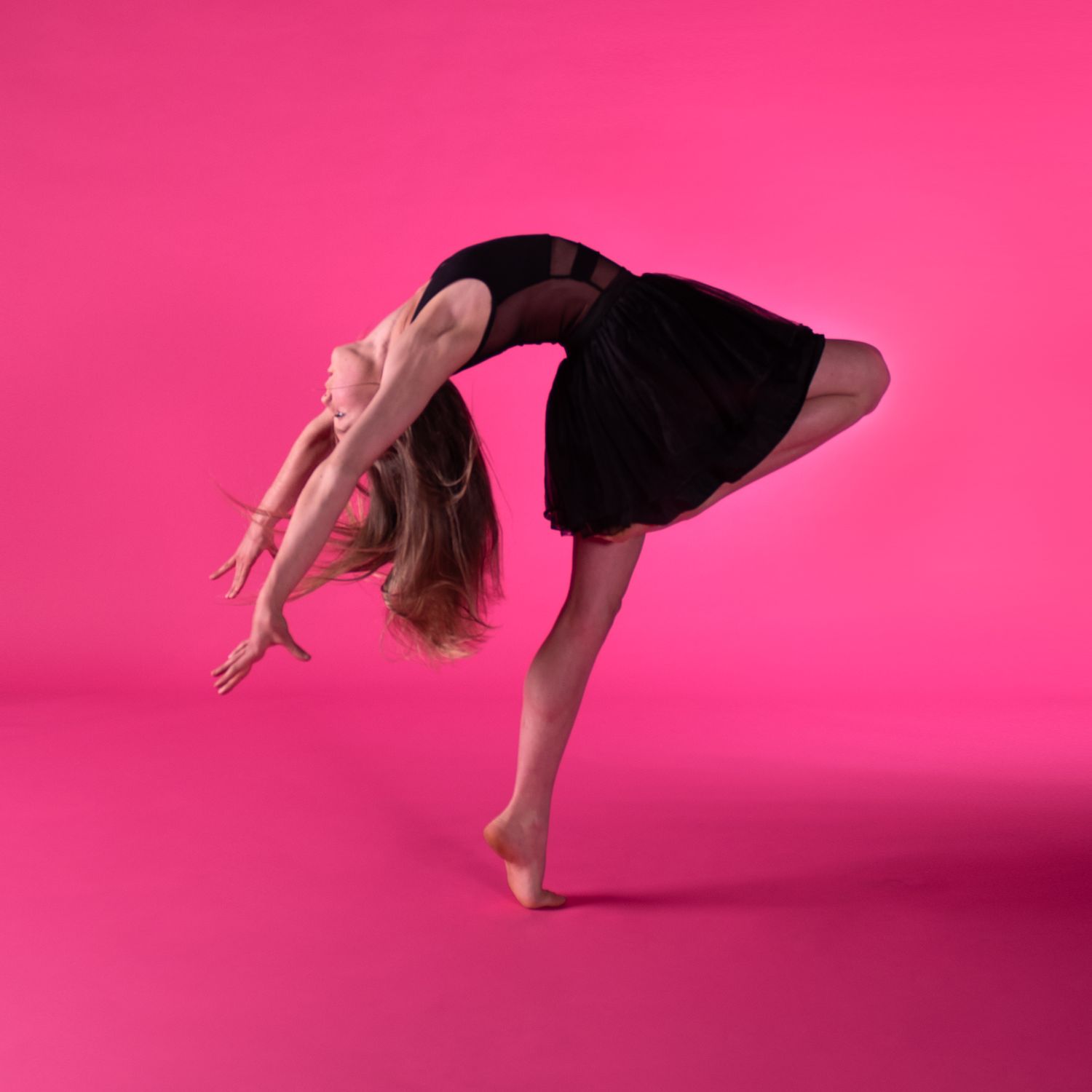 A dancer in a black dress performs a graceful backward bend pose on a bright pink background.