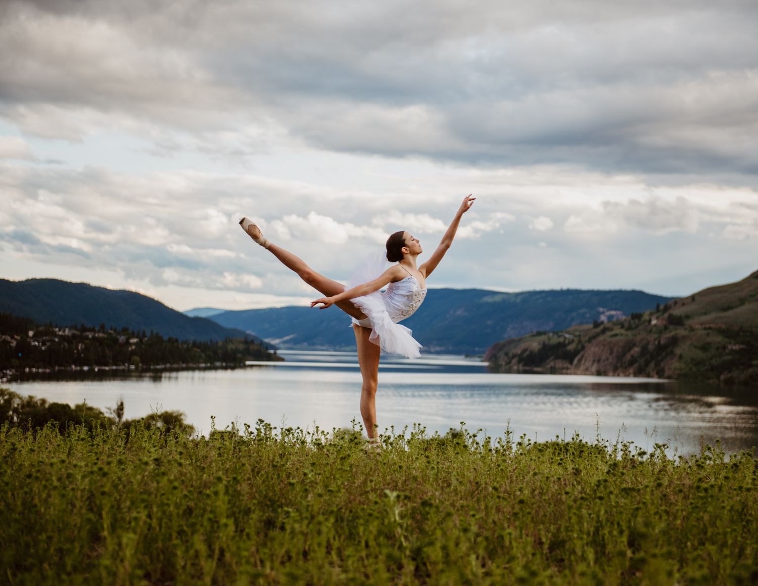 A dancer performs an elegant arabesque pose in a grassy meadow overlooking a serene mountain lake on a cloudy day.