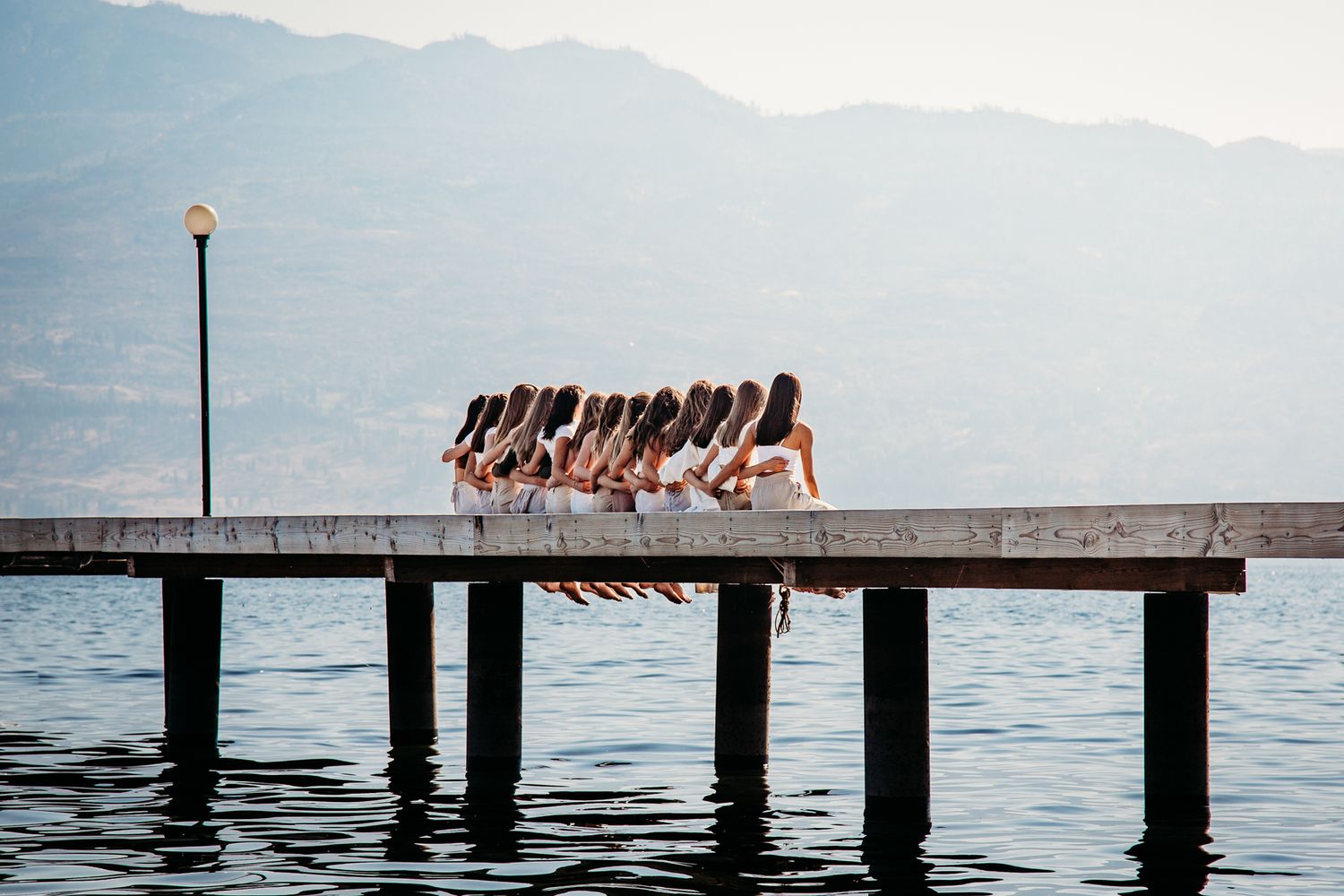 A group of dancers in dark attire perform in formation on a wooden pier over a tranquil lake with mountains in background.