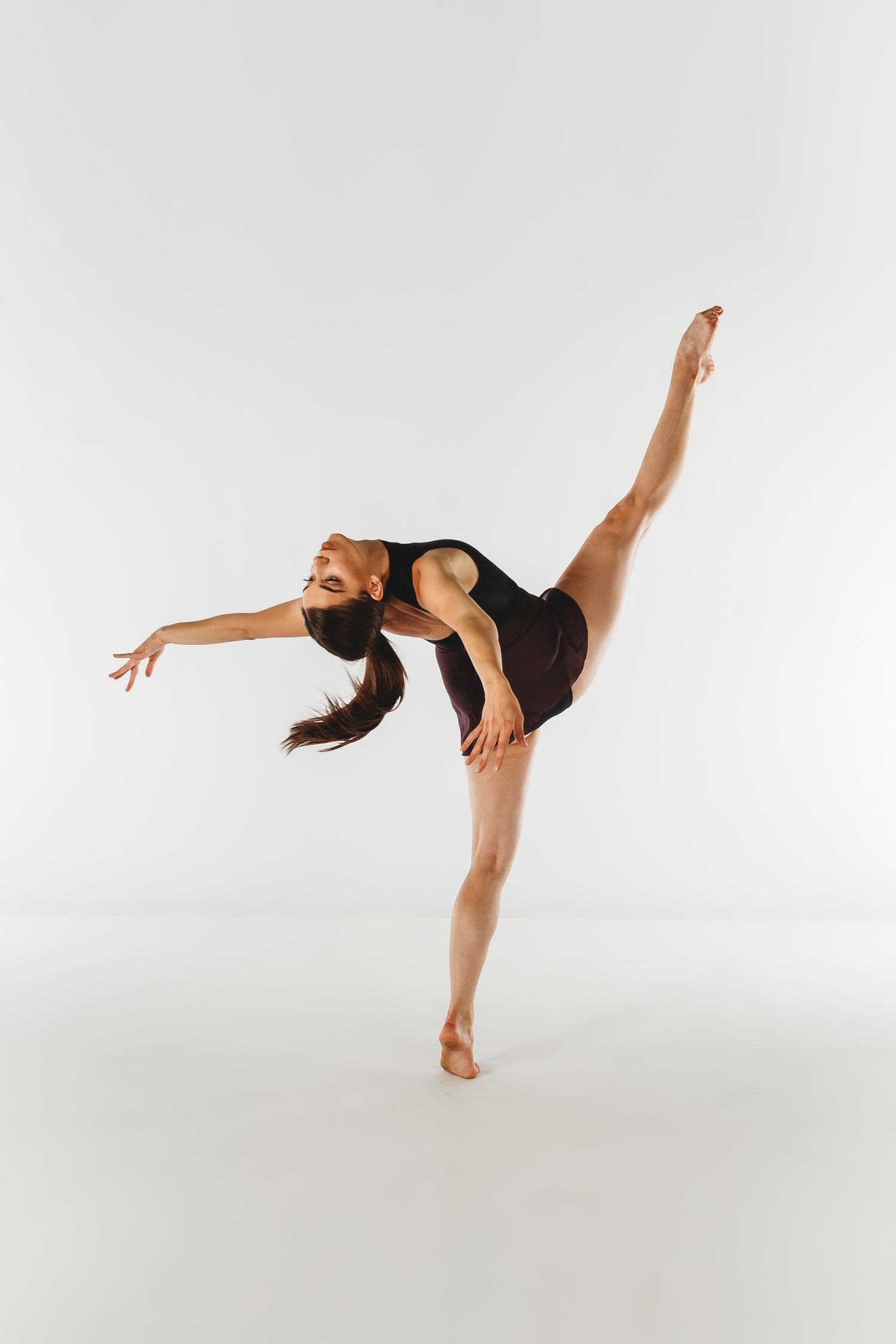 Modern dancer in black leotard performing an elegant arabesque pose against white background.