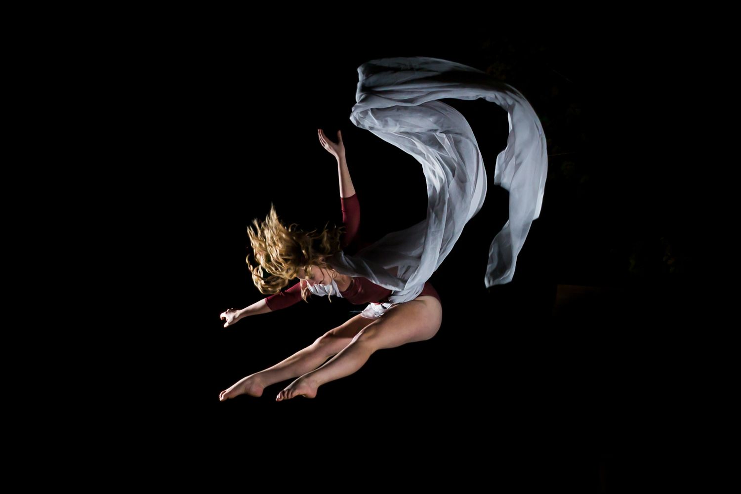 Dancer in flowing white fabric performs graceful arching movement against black background.