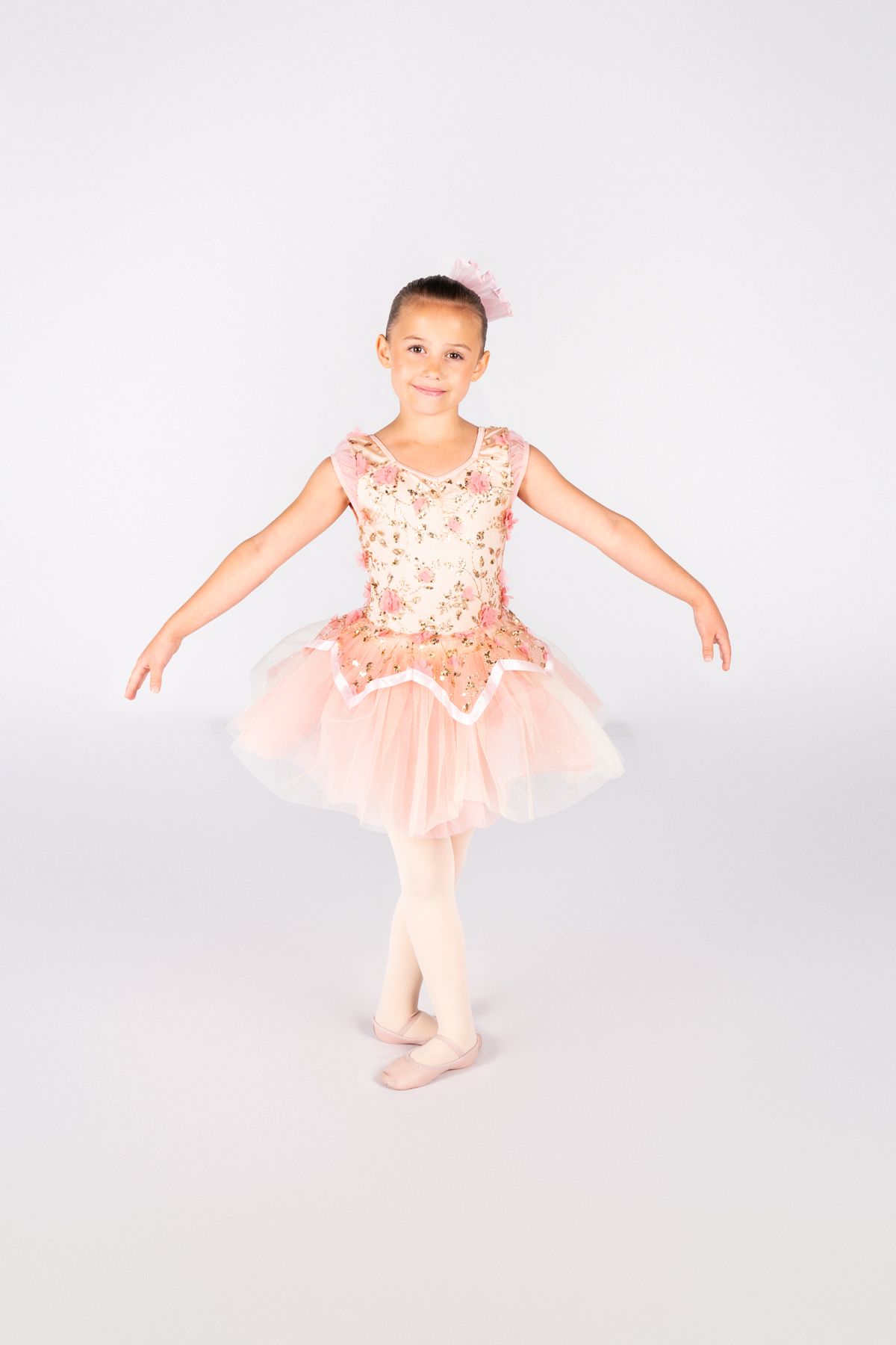 Young ballet dancer in peach sequined tutu strikes a graceful pose against white background.