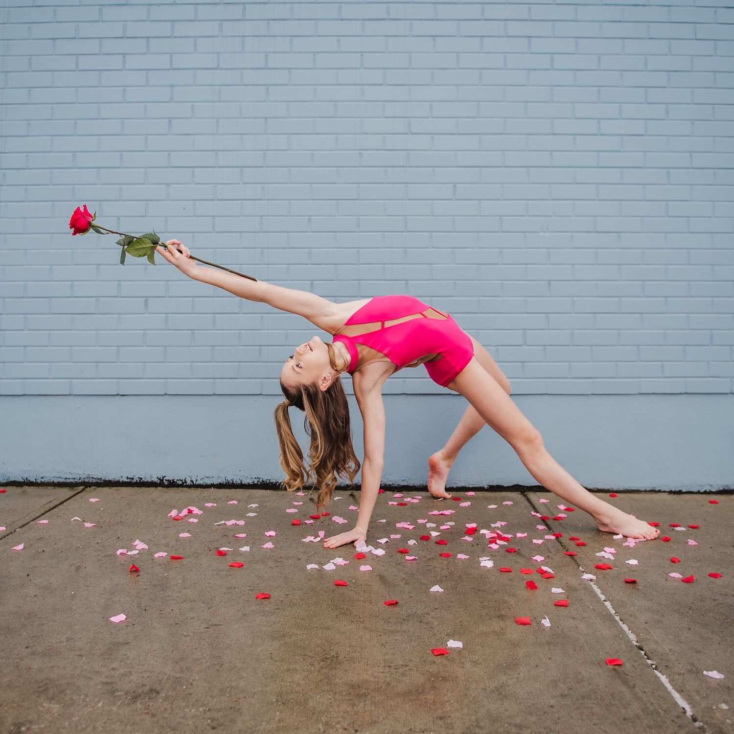 Dancer in pink leotard performing backbend with rose petals scattered on ground against grey wall.