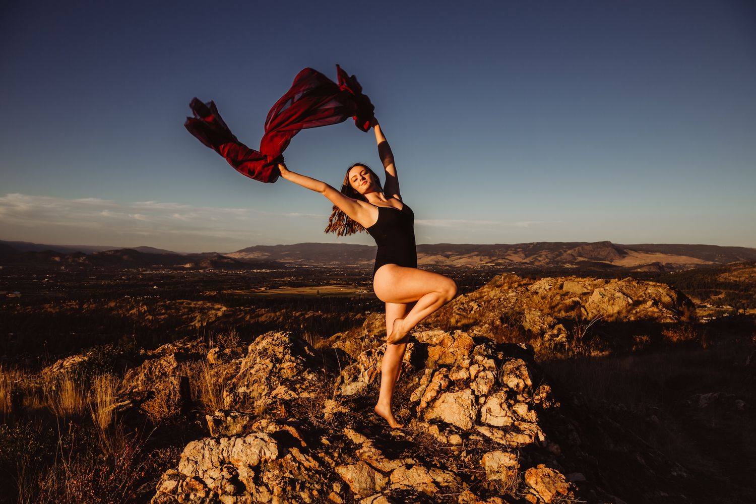 A dancer leaps gracefully with flowing red fabric on a rocky mountain overlook at dusk.