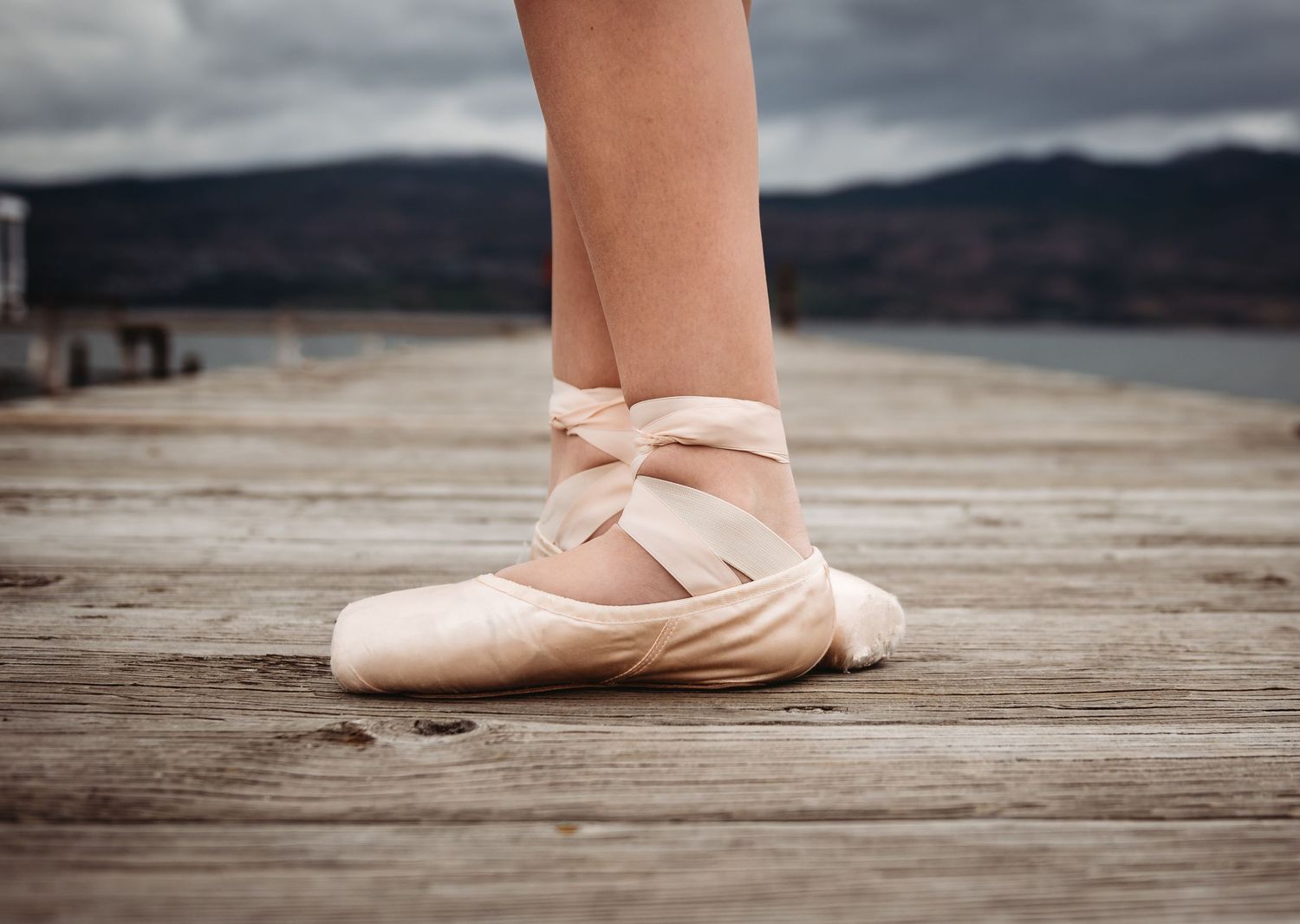 A ballet pointe shoe rests on a wooden dock with mountains in the distance under stormy skies.