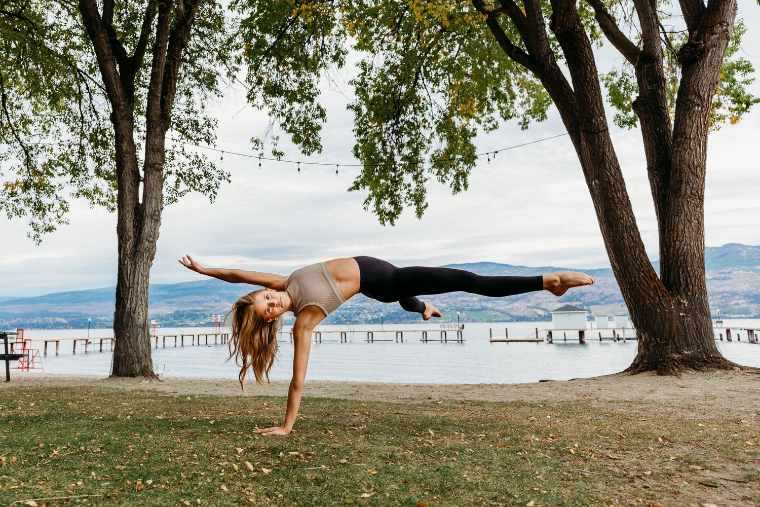 A gymnast performs a mid-air split between two trees with a scenic mountain lake view in the background.