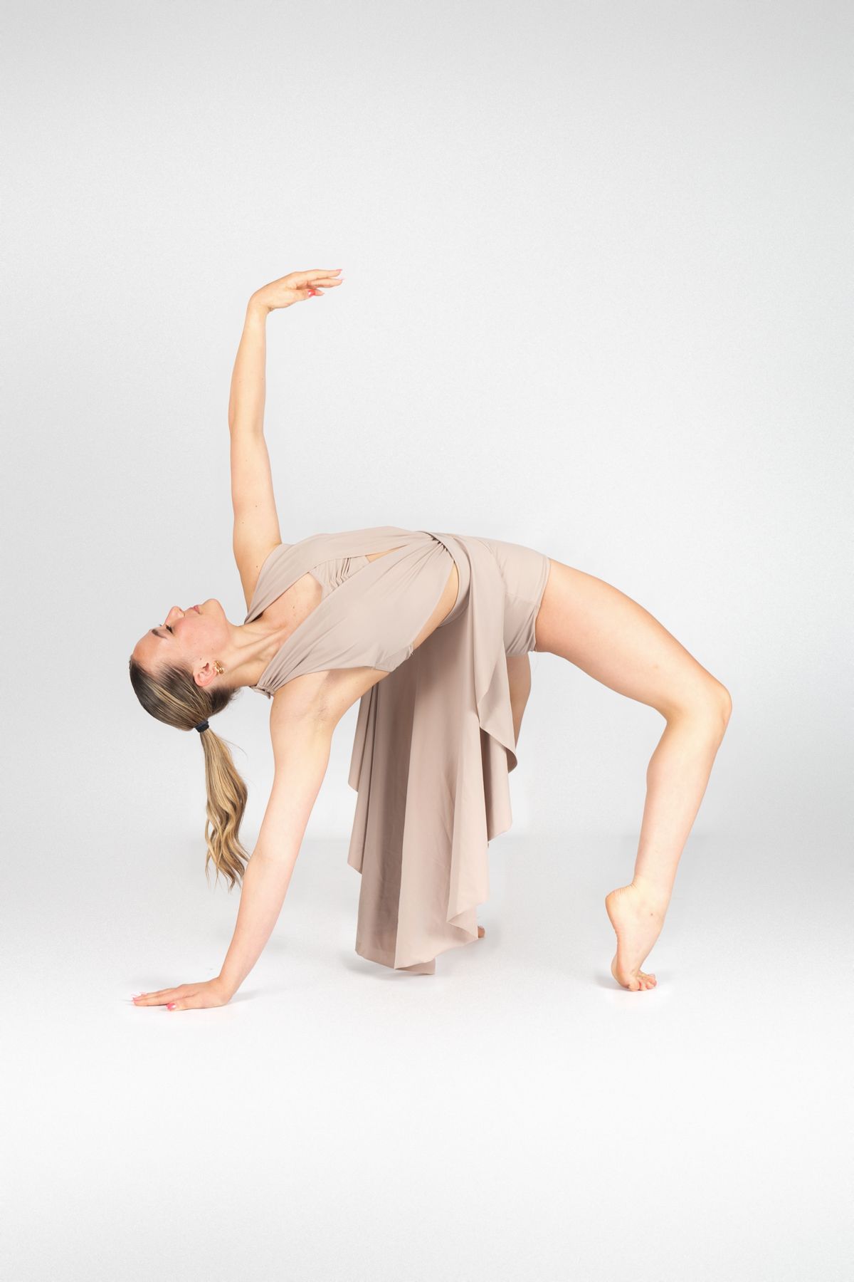 Dancer in beige dress performs graceful backbend pose against light gray background in studio.